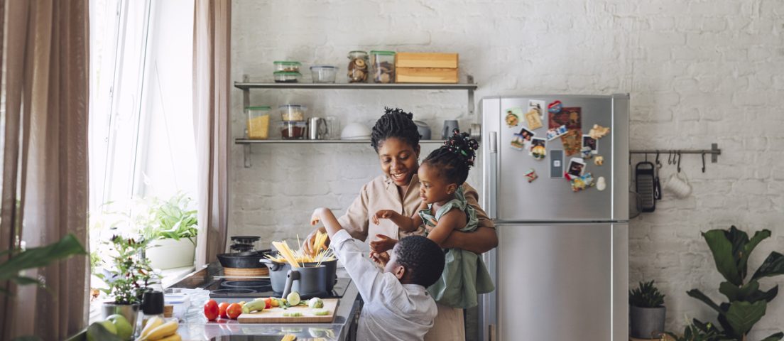 Happy African American mother, daughter and son preparing spaghetti and vegetables for lunch over a kitchen sink.